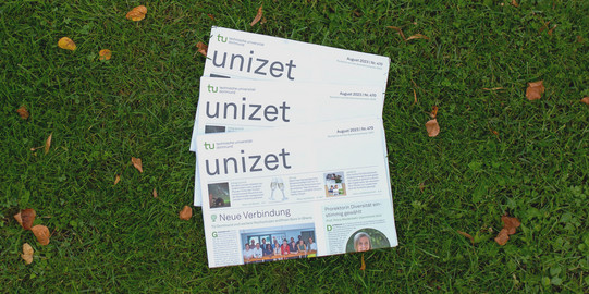 Three issues of unizet from August 2023 lie fanned out on top of each other in the grass.