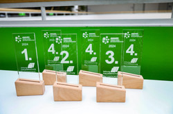 Auf dem Bild sind 6 Preise aus Glas mit Holzsockel für die Preisverleihung Junge Spitzenforschung auf einem weißen Tisch mit grünem Hintergrund zu sehen.
