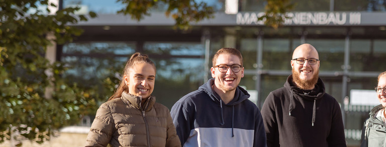 Vier Auszubildende stehen draußen vor dem Maschinenbaugebäude der TU Dortmund.
