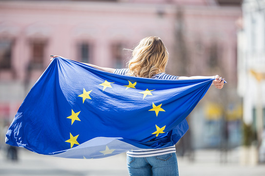 A person with long blond curls is carrying a European flag stretched behind him.