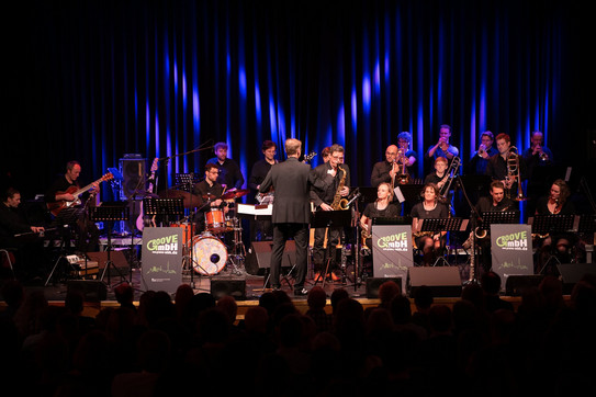 A large jazz ensemble perfomed on a stage. A conductor stands with his back to the camera and leads the band.