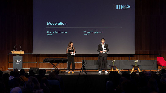 Zwei Personen stehen auf einer Bühne und moderieren die Veranstaltung