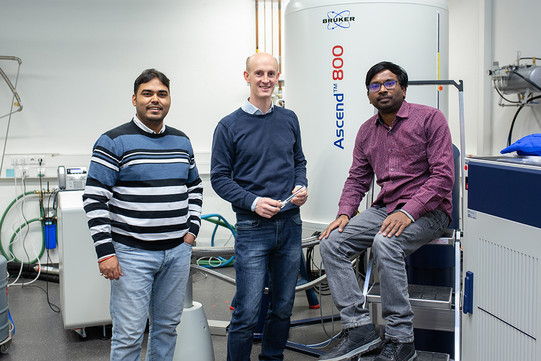Drei Forscher in Pullovern sitzen vor einem NMR-Spektrometer.