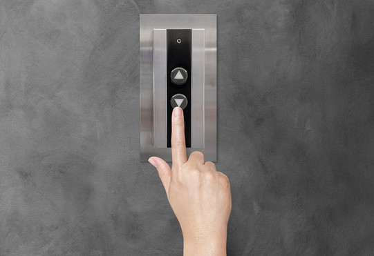 Eine Hand drückt den Abwärtsknopf vor einem Aufzug.