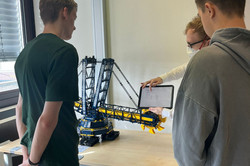 Mitarbeiter erklärt Teilnehmenden etwas auf einem Tablet. Im Hintergrund ein Modell einer Beförderungsmaschine.