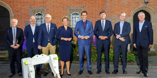 A woman and seven men stand side by side for a group photo outside a building.