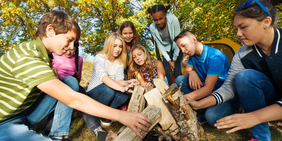 In the picture, eight young people are squatting around a small wooden tipi in the forest