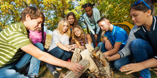 In the picture, eight young people are squatting around a small wooden tipi in the forest