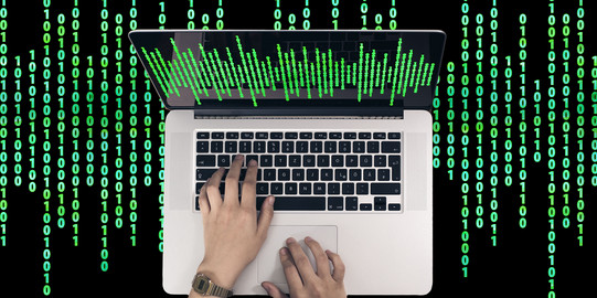 Two hands typing on the keyboard of a laptop. In the background are green zeros and ones on black background.