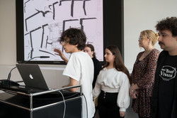 Ein Student mit braunen Haaren und Brille steht an einem Rednerpult und hält eine Präsentation. Hinter ihm stehen drei Studentinnen und ein Student.