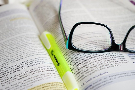 In Nahaufnahme ist ein aufgeschlagenes Buch zu sehen, auf diesem liegen Stift und Brille.
