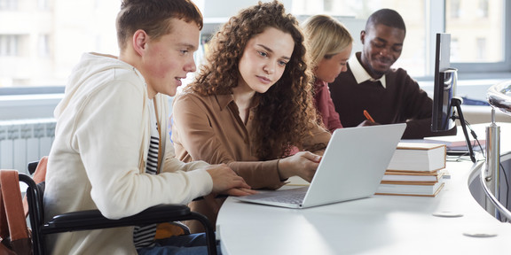 Das Bild zeigt junge Menschen mit körperlicher Beeinträchtigung und ohne Beeinträchtigung an einem Tisch sitzen