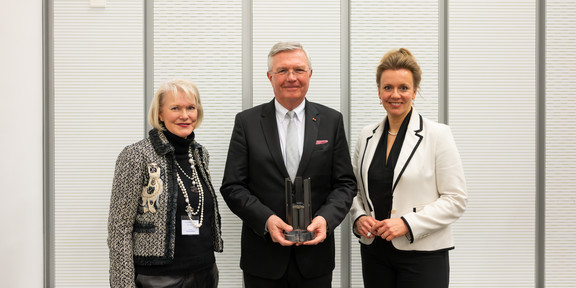 A man wearing a suit stands in the center and holds an award in his hands. To his left stands an elderly lady wearing many pearl necklaces and formal clothing. To his right, a woman wearing a white blazer and a black dress.