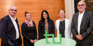 Zwei Männer und drei Frauen stehen um einen Stehtisch. Auf dem Stehtisch steht ein grüner TU-Logo-Aufsteller.