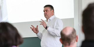 The picture shows Chief Sales Officer and TU alumnus Dr. Jan Dienstuhl in a white long-sleeved shirt in front of a screen