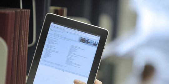 A hand holds a tablet on a bookshelf in the university library.