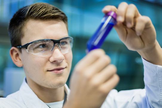 A male student wearing safety glasses holds a test tube with a blue liquid in his hands.