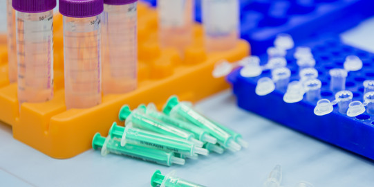 Mehrere Probenbehälter stehen in einem Fach, Zentrifugengefäße und Spritzen liegen daneben im Labor.