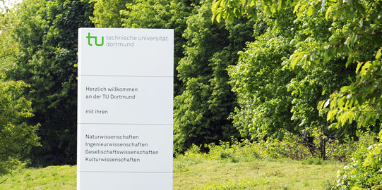 A white welcome information board of the TU Dortmund University stands on a green lawn.