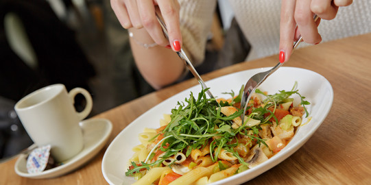 A plate with pasta and a cup are standing on a table. Hands are cutting the food.