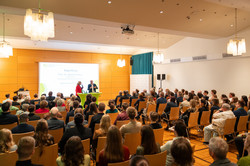Das Bild zeigt die Publikumsreihen im Saal des Westfälischen Industrieklubs von hinten mit Blick auf die Bühne, wo sich Rektor Manfred Bayer und Moderatorin Britt Lorenzen gerade unterhalten.
