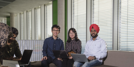 Internationale Studierendengruppe_059 Zwei junge Männer, einer asiatisch aussehend, der andere mit einem Turban, und eine asiatisch aussehende junge Frau sitzen in einem Gebäude auf einem Sofa mit Laptops auf dem Schoß und lächeln in die Kamera.