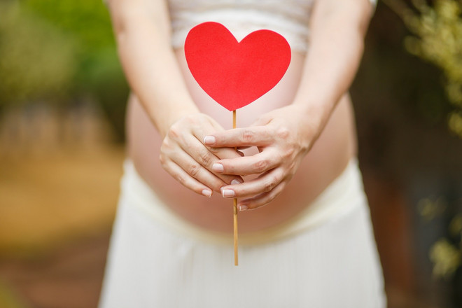 Eine schwangere Person hält ein gebasteltes rotes Herz an einem Holzstiel vor den Bauch.