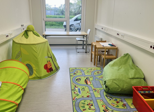 Es ist ein Raum gezeigt, Der am Ende eine Fensterfront hat Mit einem Ausblick auf Grüne Sträucher. Auf dem Boden des Raumes ist ein Kinderspielteppich mit bunten Straßen Punkt auf diesem Teppich befindet sich ein Sitzsack und eine Kisten Kinderspielzeug in Des Weiteren befindet sich auf der linken Seite ein Tunnelschlauch zum Durchkrabbeln für kleine Kinder und ein Kinderzelt. Im hinteren Teil des Raumes ist ein Kindertisch mit Kinderstühlen und ein weiterer Stuhl.