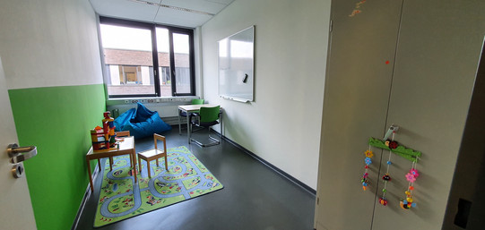 Bild aufgenommen durch geöffnete Tür, zeigt Eltern-Kind-Raum, Teppich mit Spielzeug, Sitzmöglichkeiten, Whiteboard an der Wand