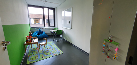 Eltern-Kind-Raum Bild aufgenommen durch geöffnete Tür, zeigt Eltern-Kind-Raum, Teppich mit Spielzeug, Sitzmöglichkeiten, Whiteboard an der Wand