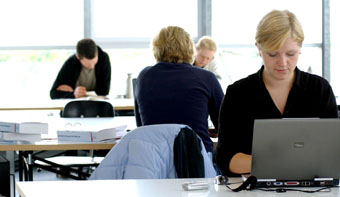 Four persons sitting at tables in front of a large window, reading or working on their computers