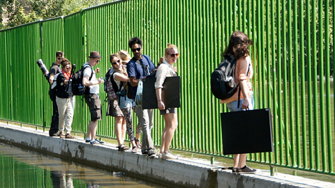 Mehrere Personen balancieren mit Ihren Kunstmappen in der Hand über einen Vorsprung vor einem Wasserbecken und halten sich dabei an einem Gitter fest