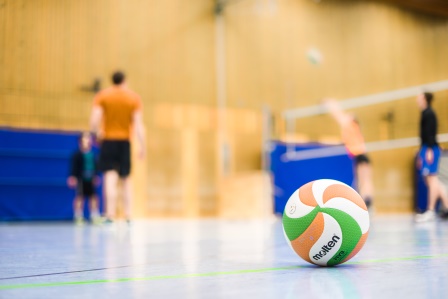 A volleyball on the floor of a sports hall. In the background, you can see a volleyball net and four people.
