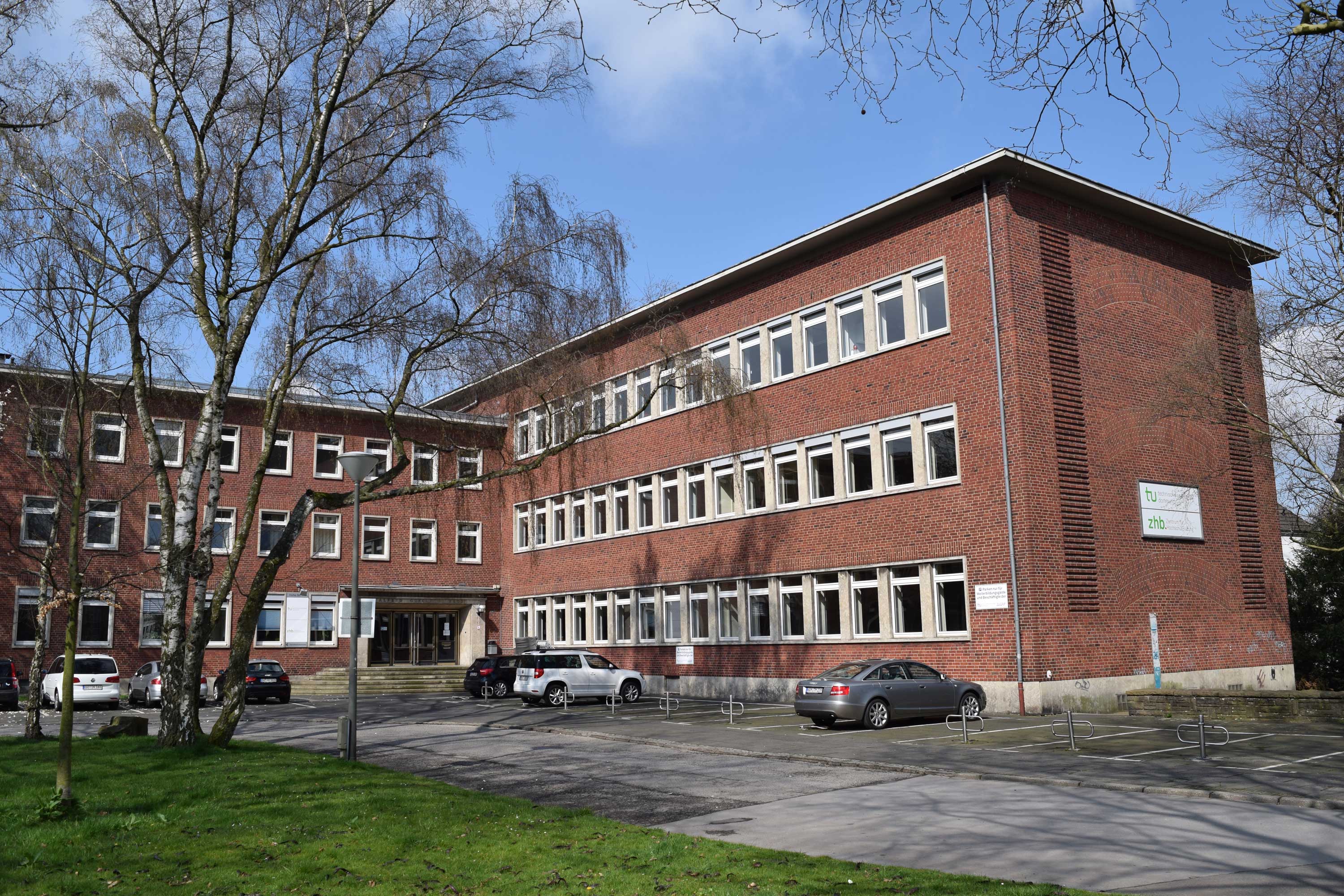 Dreistöckiges Backsteingebäude mit Bäumen und Grün im Vordergrund