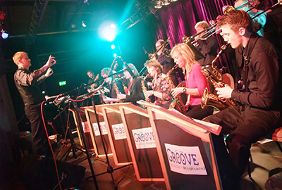 Big Band Groove m.b.H. A conductor conducts the musicians. The name Groove m.b.H. is written on the music stands.