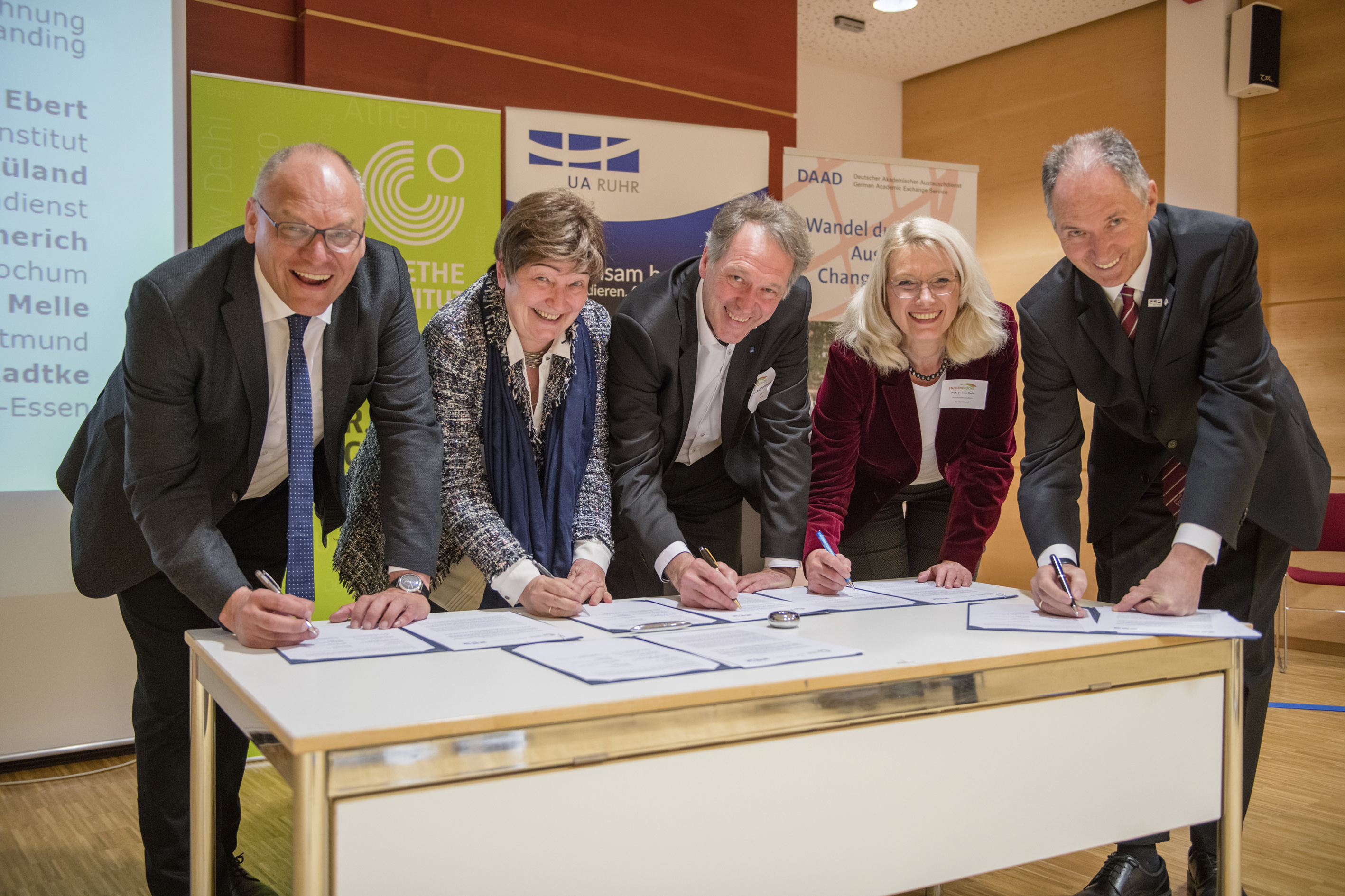 Studienbrücke Fünf Personen stützen sich neben einander auf einen Tisch und unterschreiben ein Dokument.