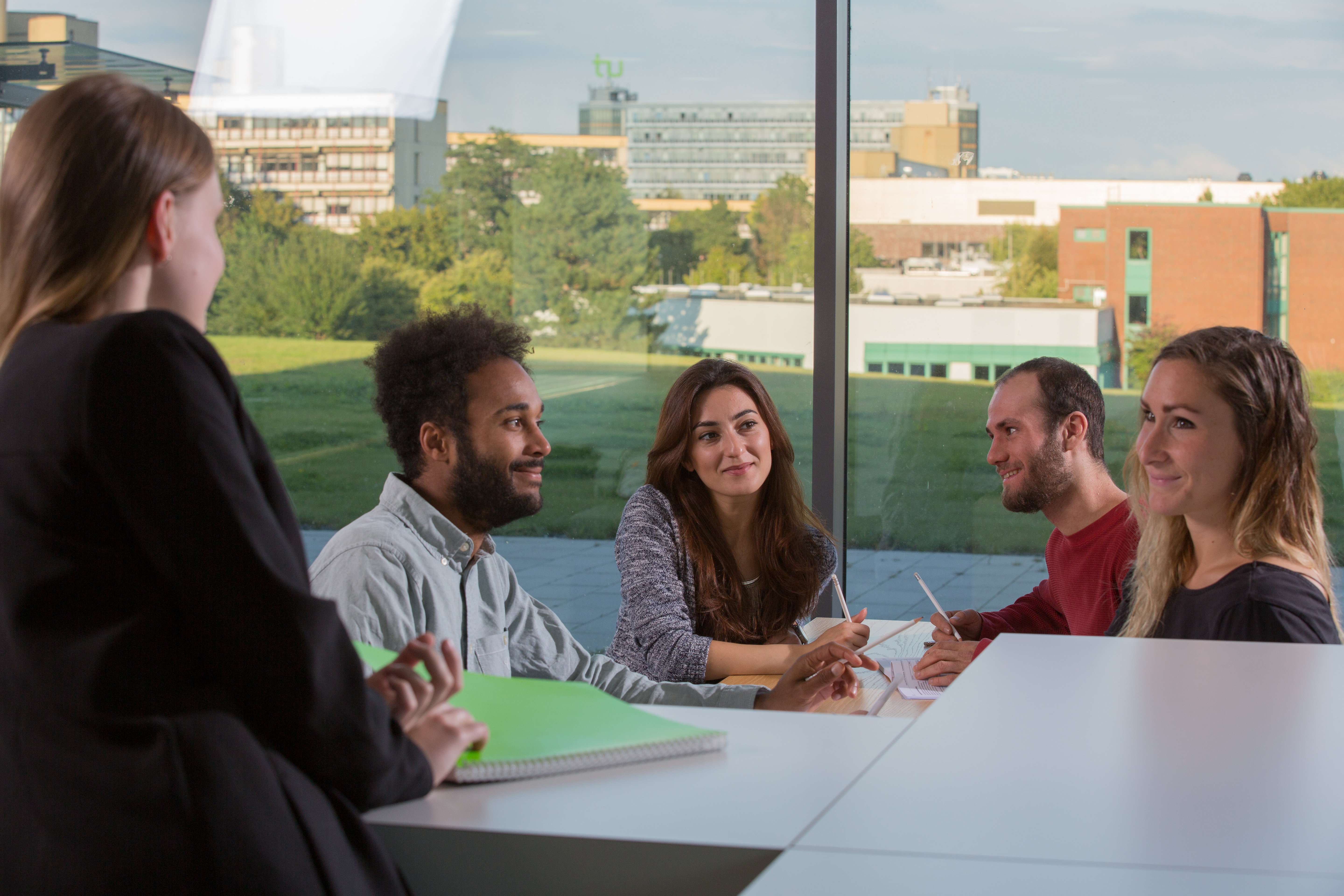 Five students sit at a table with pens in their hands and paper in front of them on the table.