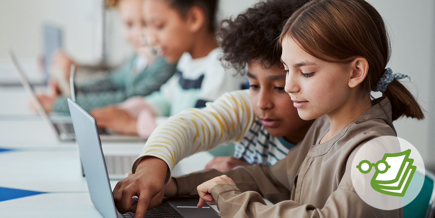 Zwei Kinder sitzen an einem Tisch und tippen auf einem Laptop. Im Hintergrund sind weitere Kinder mit Laptops an Tischen zu sehen.