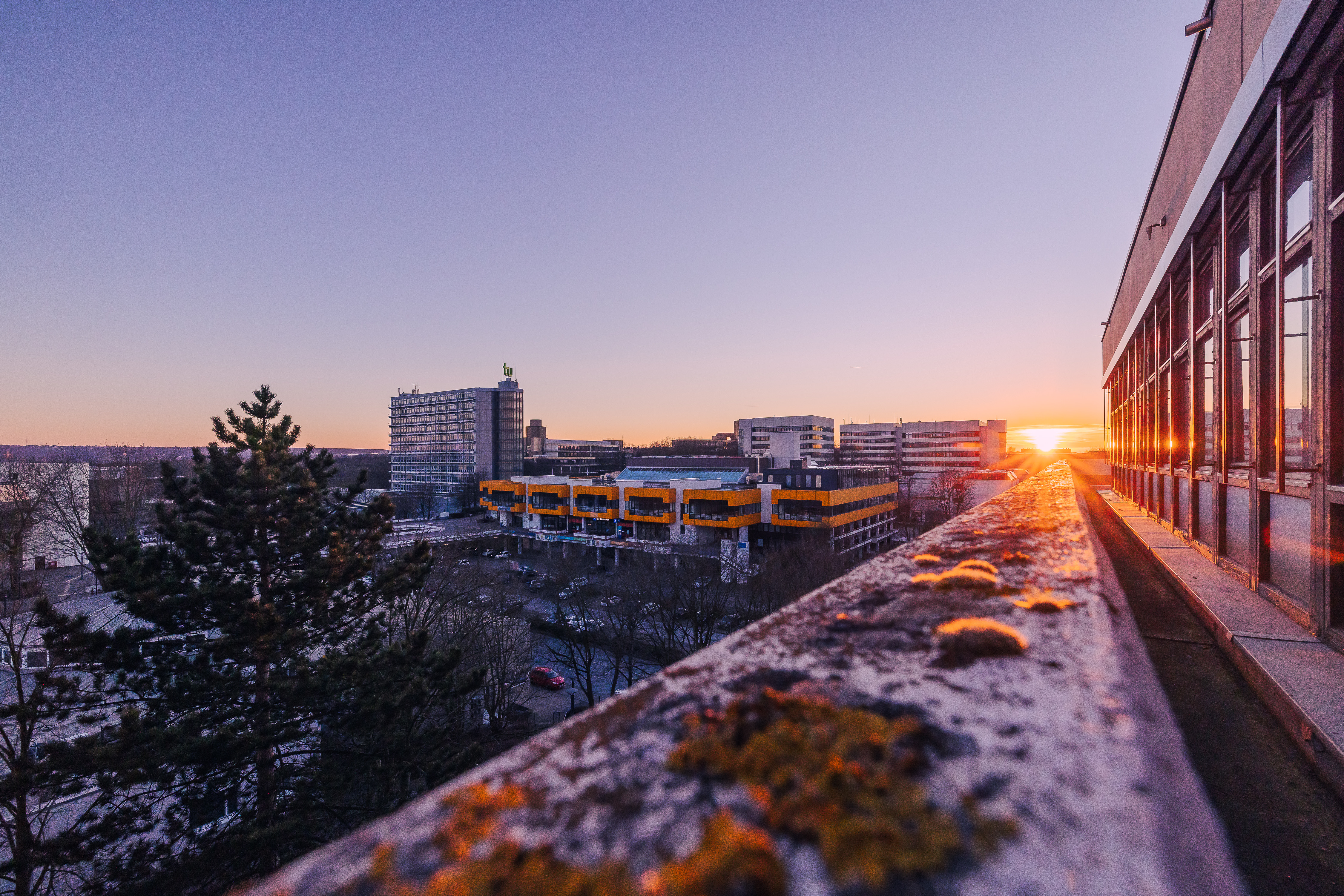 Der vereiste Campus der TU Dortmund mit dem Mathetower und dem Mensagebäude im Hintergrund sind vom eisbedeckten Balkon eines Gebäudes zu sehen. Im Hintergrund geht die Sonne unter.