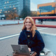 Eine Frau sitzt im Freien auf einem Stuhl mit einem Laptop und lächelt in die Kamera.