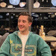 Ein Student sitzt in einer Gastronomie in einer blauen Jacke und grinst leicht nach rechts. 
