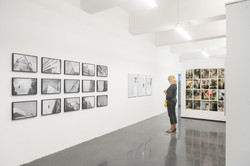 Eine Frau liest ein Essay an einer Ausstellungswand. Links vorne sind 15 schwarz-weiß Fotografien in drei Fünfer-Reihen angeordnet.