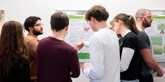 Im Zentrum des Bildes sind vier junge Erwachsene seitlich und von hinten zu sehen, die gemeinsam ein Plakat anschauen, welches an einer weißen Wand befestigt ist. Links im Vordergrund ist eine Person mit schwarzem Oberteil und braunen Haaren, ebenfalls von Hinten zu sehen. Rechts von den Personen steht eine männlich gelesene Person seitlich vor einem weiteren Plakat und steht mit dem Rücken zur anderen Gruppe.
