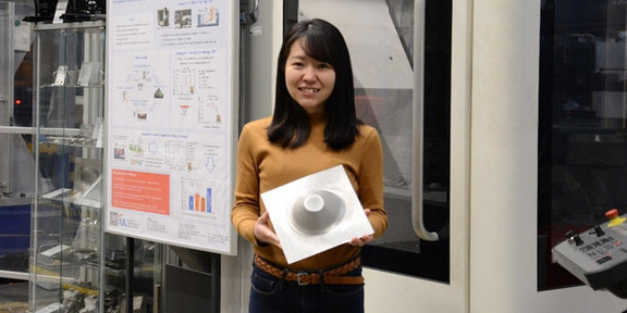 Protrait Dr. Shiori Gondo Eine junge Frau aus Japan hält ein geformtes Metalblech vor Maschinen in ihren Händen und lächelt in die Kamera.