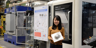 A young woman from Japan holds a shaped metal sheet in front of machines in her hands and smiles at the camera.