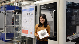 Eine junge Frau aus Japan hält ein geformtes Metalblech vor Maschinen in ihren Händen und lächelt in die Kamera.