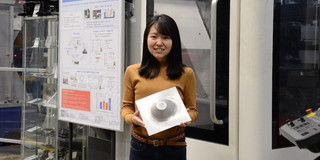 Protrait Dr. Shiori Gondo Eine junge Frau aus Japan hält ein geformtes Metalblech vor Maschinen in ihren Händen und lächelt in die Kamera.