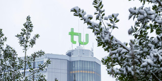 Im Vordergrund schneebedeckte Zweige, im Hintergrund der Mathetower mit grünem TU-Logo auf dem Dach
