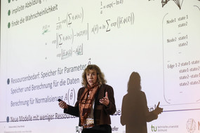 Eine Frau erklärt etwas, in der Präsentation im Hintergrund sind mathematische Formeln zu sehen