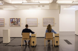 Zwei Personen sitzen in Shaker-Stühlen vor Bildern in einem Ausstellungsraum.